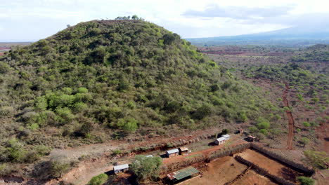 Aerial-of-fill-covered-in-green-trees-and-bushes-with-small-buildings-at-foot-of-hill-in-rural-Kenya