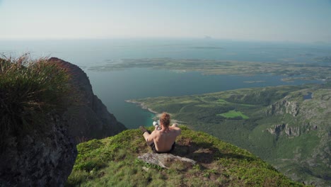 Shirtless-Man-Hiker-Sitting-On-A-Rocky-Ledge-Taking-Travel-Photos-Using-A-Long-Lens-Camera-At-Mount-Donnamannen,-Nordland,-Norway