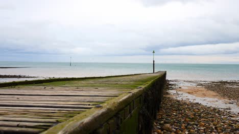 Weathered-wooden-boardwalk-jetty-leading-towards-calm-overcast-ocean-slow-descending-shot