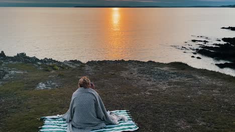 Aerial-of-young-hipster-woman-sitting-on-blanket-in-front-of-Atlantic-Ocean-in-France,-Brittany-taking-photo-during-sunset-golden-hour