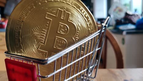 Golden-bitcoin-crypto-currency-coins-in-tiny-shopping-trolley-on-kitchen-table-concept-close-orbit-right