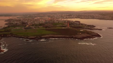 Drone-rotate-around-old-lighthouse-in-la-coruna-city-of-north-Spain-Galicia-region,-aerial-footage-at-sunset-cityscape-harbor-and-Atlantic-Ocean-beach-rock-cliff-formation