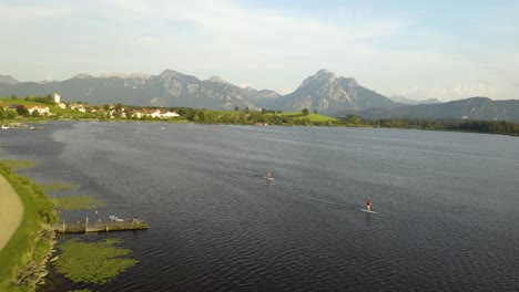 Aerial-View-of-People-Using-Stand-Up-Paddle-Boards-on-Lake-in-Bavaria,-Mountains-in-Background