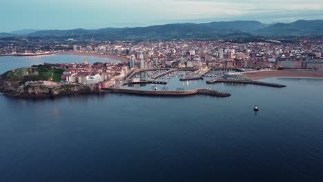 Luftaufnahmen-Des-Stadtbildes-Von-Gijon,-Ozeanstrand,-Hafen-Und-Urbaner-Skyline,-Die-In-Der-Dämmerung-Nach-Sonnenuntergang-Beleuchtet-Werden