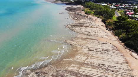 A-gorgeous-light-blue-and-green-ocean-pushes-its-small-waves-into-a-shoreline-made-stone-and-rock-whilsts-the-treeline-behind-makes-for-a-hidden-beach-on-the-sunny-day-at-Hervey-Bay,-Australia