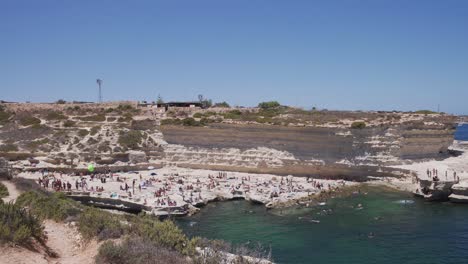 Panoramic-shot-of-St-Peters-Pool,-people-are-enjoying-and-jumping-into-the-sea-water-in-Malta