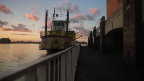 Steamboat-ferry-docked-on-water-at-sunset-with-beautiful-clouds-in-Mobile,-Alabama-at-port-panning-right-slow-motion-in-4k-with-fence-in-foreground