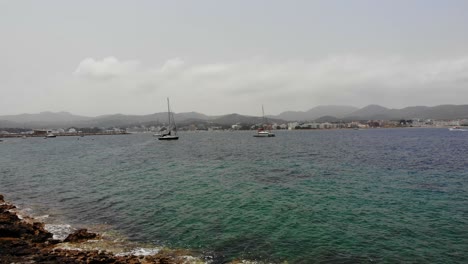Schöne-Luftaufnahme-Des-Kalksteinstrandes-In-Ibiza,-Sant-Antoni-In-Richtung-Yachten,-Die-An-Einem-Warmen-Sonnigen-Tag-Im-Kristallklaren-Smaragdblauen-Meer-Mit-Bergen-Im-Hintergrund-In-Spanien-Schwanken