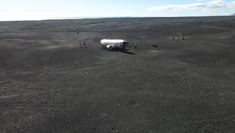People-exploring-crashed-plane-on-back-beach-of-Iceland,-aerial-fly-forward