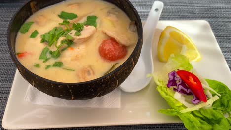 Spicy-Thai-coconut-soup-with-chicken-and-vegetables-in-a-coconut-bowl,-hot-dish-in-Thai-cuisine-restaurant,-4K-tilting-up