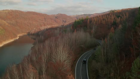 An-aerial-drone-view-shot-following-a-car-driving-on-a-winding-road-alongside-a-lake-and-a-water-dam-in-late-autumn