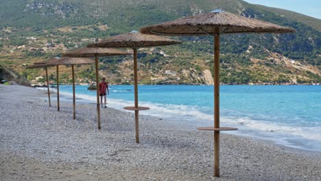 A-Male-Tourist-Relaxing-Walking-By-The-Beautiful-Shore-With-Native-Umbrellas-And-Mountains-Background-At-Agia-Kiriaki-Beach-Near-Zola-Village,-Kefalonia,-Greece