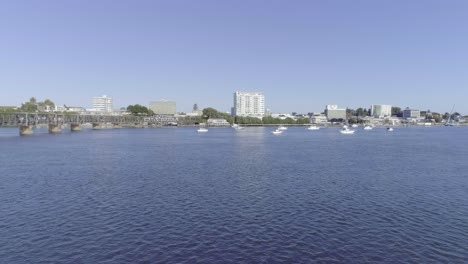 Tauranga-City-skyline-seen-from-calm-bay-of-plenty-with-boats-anchored,-sunny-day,-aerial