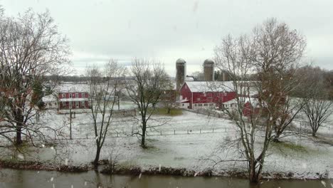 Aerial-establishing-shot-of-family-farm-with-red-barn,-brick-house-during-winter-snowstorm