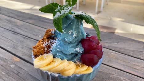 Delicious-blue-spirulina-frozen-fruit-bowl-with-bananas,-homemade-cookie-crumble,-grapes-and-mint-on-a-wooden-table,-4K-shot