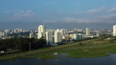 Aerial-drone-left-trucking-and-panning-shot-of-the-south-of-São-Paulo,-Brazil-from-the-man-made-Guarapiranga-Reservoir-towards-the-Socorro-neighborhood-with-skyscrapers-and-houses-on-a-fall-evening