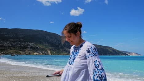 Charming-Woman-With-Smartphone-In-Hand-Walking-Away-From-The-Shore-Smiling-In-Agia-Kiriaki-Beach,-Zola-In-Kefalonia,-Greece