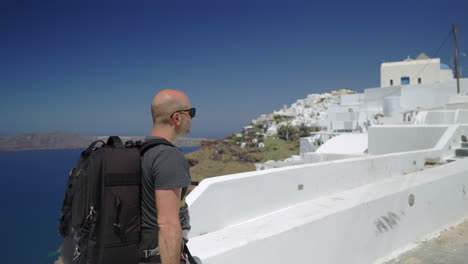 Following-from-the-side-a-man-walking-in-Santorini,-Greece,-wearing-a-backpack
