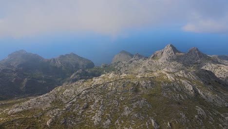 Flying-over-rigged-mountains-with-vegetation-and-the-sea-in-the-background-with-clouds-above-at-Sa-Calobra,-Mallorca,-Spain