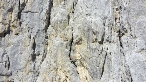 Aerial-backwards-shot-of-mountaineer-climbing-on-steep-cliff-wall-during-sunny-day---Marmolada,-Dolomites