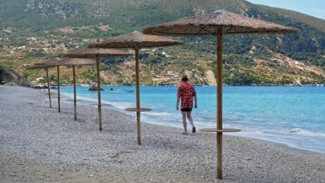A-Man-In-Summer-Beach-Outfit-Walking-Along-The-Shoreline-Pebbled-Beach-Of-Agia-Kiriaki,-Zola-In-Kefalonia,-Greece
