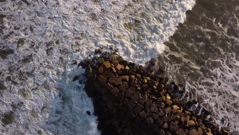 Aerial-top-down-shot-of-stormy-waves-of-ocean-crashing-against-rocky-jetty-during-sunset---Circle-shot