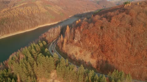 An-aerial-descending-drone-view-of-a-car-driving-on-a-winding-road-alongside-a-lake-in-late-autumn