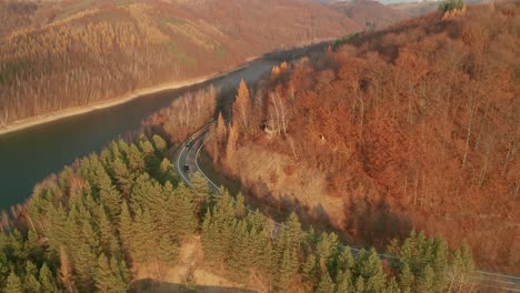 An-aerial-ascending-drone-view-of-two-cars-driving-on-a-winding-road-alongside-a-lake-in-late-autumn