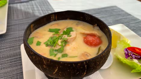 Würzige-Thailändische-Kokosnusssuppe-Mit-Hühnchen-Und-Gemüse-In-Einer-Kokosnussschüssel,-Warmes-Gericht-Im-Thailändischen-Küchenrestaurant,-4k-aufnahme