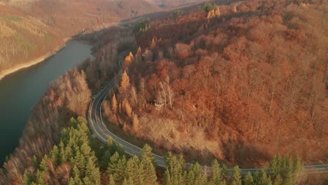 An-aerial-ascending-drone-view-of-a-car-driving-on-a-winding-mountain-road-alongside-a-lake-in-late-autumn