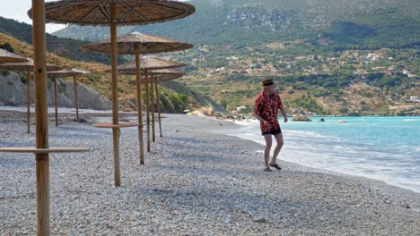 Man-Wearing-Hat-And-Summer-Beach-Floral-Outfit-Throwing-Pebbles-Into-The-Sea-In-Agia-Kiriaki-Beach,-Zola,-Kefalonia,-Greece