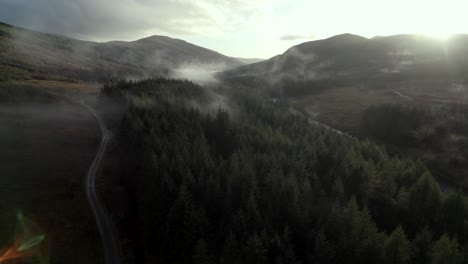 Aerial-drone-footage-slowly-descending-into-a-dark-forest-of-conifer-trees,-a-road-and-a-river-while-shafts-of-light-illuminate-low-hanging-cloud-in-the-treetops-at-sunset