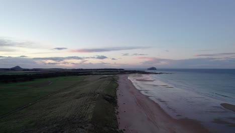 Drone-footage-flying-slowly-backwards-over-a-long-sandy-beach-and-sand-dunes-during-a-pink-sunset-as-the-tide-gently-laps-the-shore-to-reveal-people-walk-along-the-beach