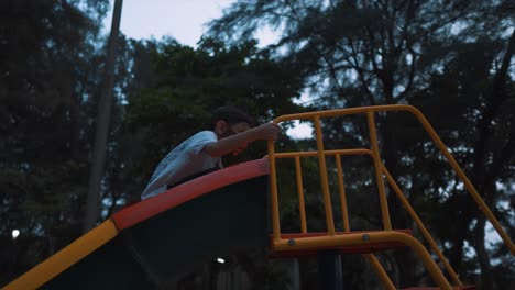 A-young-Asian-boy-playing-alone-on-a-park-slide-having-fun-as-he-slides-backwards-down-the-slide-as-the-sun-sets-in-the-evening-on-a-beautiful-day-outdoors