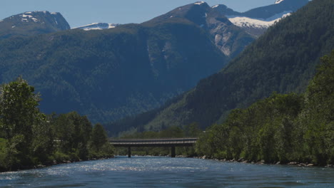 Kristallblauer-Fluss,-Der-Auf-Eine-Brücke-Vor-Hohen,-Schneebedeckten-Bergen-Zufließt