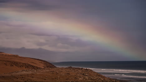 Time-lapse-of-huge-rainbow-over-coastal-rocky-coast-while-waves-breaks-moon-shore