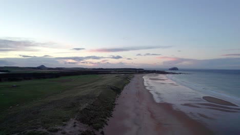 Drone-footage-flying-over-a-long-sandy-beach-and-sand-dunes-during-a-pink-sunset-as-the-tide-gently-laps-the-shore-and-people-walk-along-the-beach