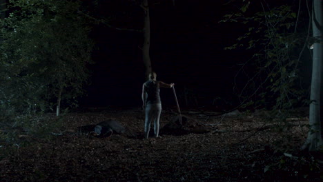 Woman-digging-a-shallow-grave-next-to-a-dead-body-in-a-spooky-forest-at-night