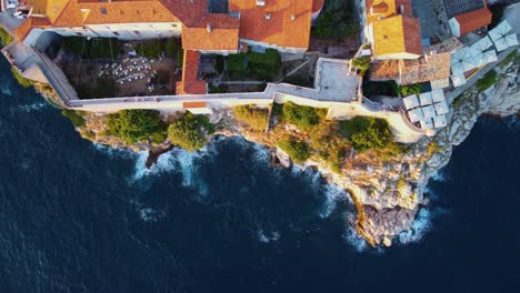 Top-down-aerial-view-in-Dubrovnik-Croatia-of-waves-crashing-into-the-cliffs-below-the-city-walls-at-sunset