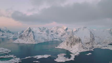Wintry-extreme-Lofoten-Reine-snow-covered-mountain-peaks-and-picturesque-blue-seascape-elevated-aerial-view