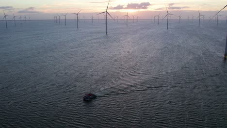 Boat-traveling-in-front-of-tall-wind-turbines-at-offshore-windpark,-dawn