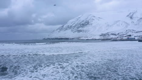 Olas-Del-Océano-ártico-E-Invierno-En-La-Ciudad-De-Olafsfjordur,-Imponentes-Montañas-Cubiertas-De-Nieve-En-El-Fondo--Norte-De-Islandia,-Vuelo-Aéreo