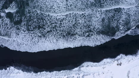 Luftaufnahme-Von-Oben-Nach-Unten-Von-Meereswellen,-Die-Den-Schwarzen-Sand-Des-Vulkanischen-Strandes-Erreichen,-Der-Mit-Schnee-Und-Eis-In-Island-Bedeckt-Ist