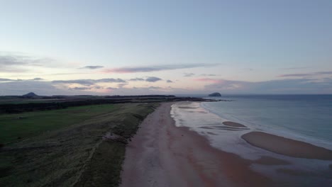 Drone-footage-flying-slowly-backwards-over-a-long-sandy-beach-and-sand-dunes-during-a-pink-sunset-as-the-tide-gently-laps-the-shore-and-people-walk-along-the-beach