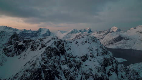 Flying-over-rugged-Lofoten-snow-covered-Svartvatnet-head-mountains-aerial-view-with-cold-sunrise-clouds