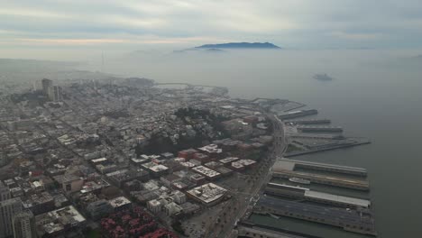 Aerial-view-of-coastal-piers-and-buildings,-foggy-evening-in-San-Francisco,-USA---pan,-drone-shot