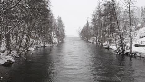 Aerial-shot-of-a-small-river-stream-by-a-lake-in-Sweden-during-snowfall-on-a-cold-winter-day