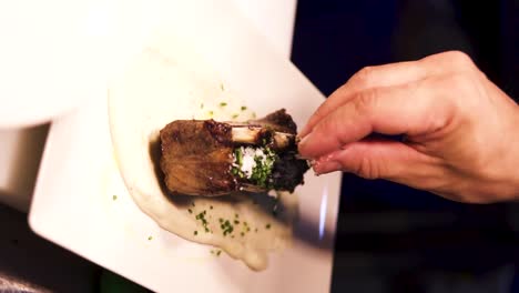 Vertical-slow-motion-shot-of-tasty-spare-ribs-garnished-with-herbs-and-lying-in-a-white-sauce-while-a-chef-adds-salt-to-the-meat-in-a-fine-restaurant