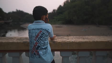 -A-young-boy-excited-climbing-up-onto-a-stone-bridge-looking-over-to-spot-fish-and-other-creatures-along-the-bank-of-the-Mandovi-River-at-sunset,-Goa,-India