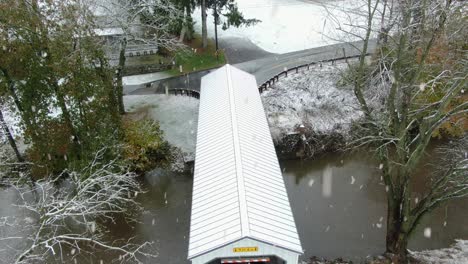 Overhead-aerial-view-of-white-covered-bridge-crossing-creek-during-winter-snow